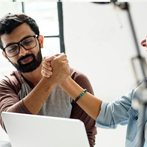 A bearded man wearing glasses clasping hands with a colleague and looking a bit uneasy.