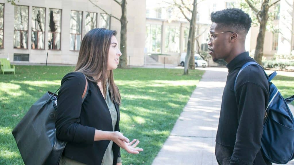 A woman wearing a bag is talking to a man wearing glasses outdoors.