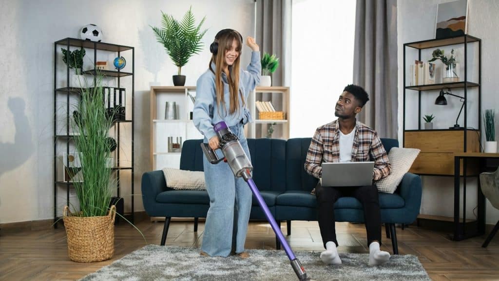 A man sitting on a couch while using his laptop looks at a woman with headphones using a vacuum cleaner.