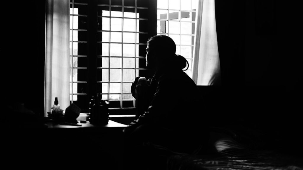 A dramatic black and white silhouette of a person sitting by a grid-patterned window, looking outside in a dark room.