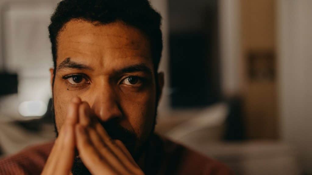 A bearded man looking distressed and holding his hands up to his face.