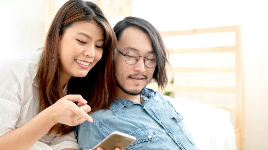 A smiling woman and a man with glasses and long hair are looking at a smartphone together.