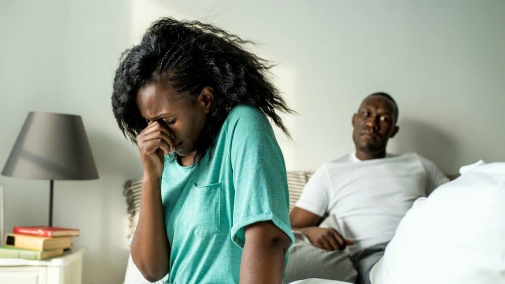 A distressed woman in a teal shirt is sitting on a bed with her hand to her face, with a displeased man sitting behind her.