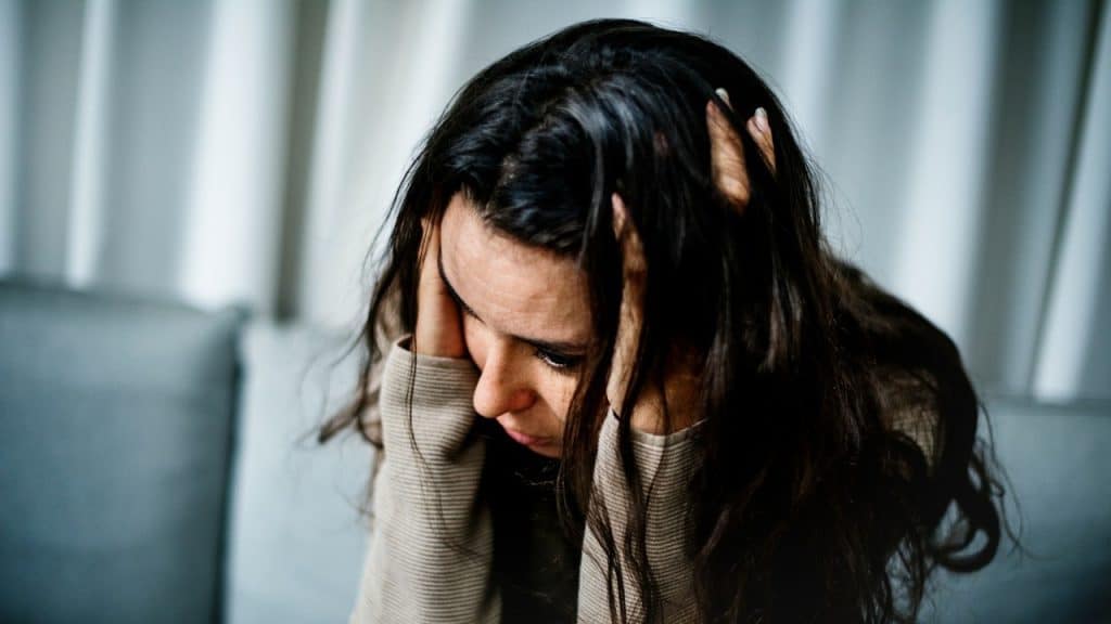 A woman with dark hair, sitting on a couch and holding her head in distress.