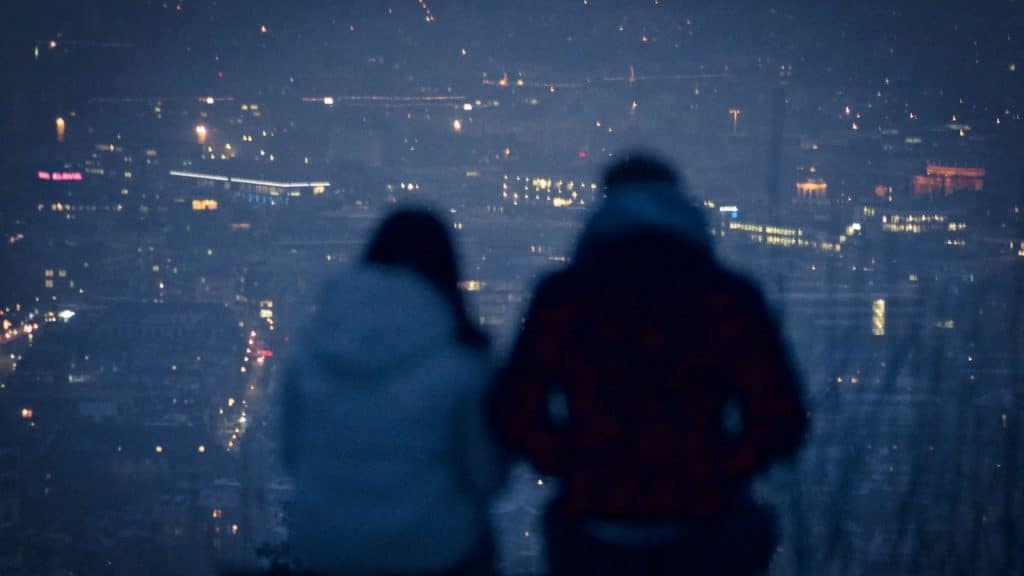 A heavily blurred, nighttime image showing the backs of two people sitting and looking out over a city with twinkling lights.