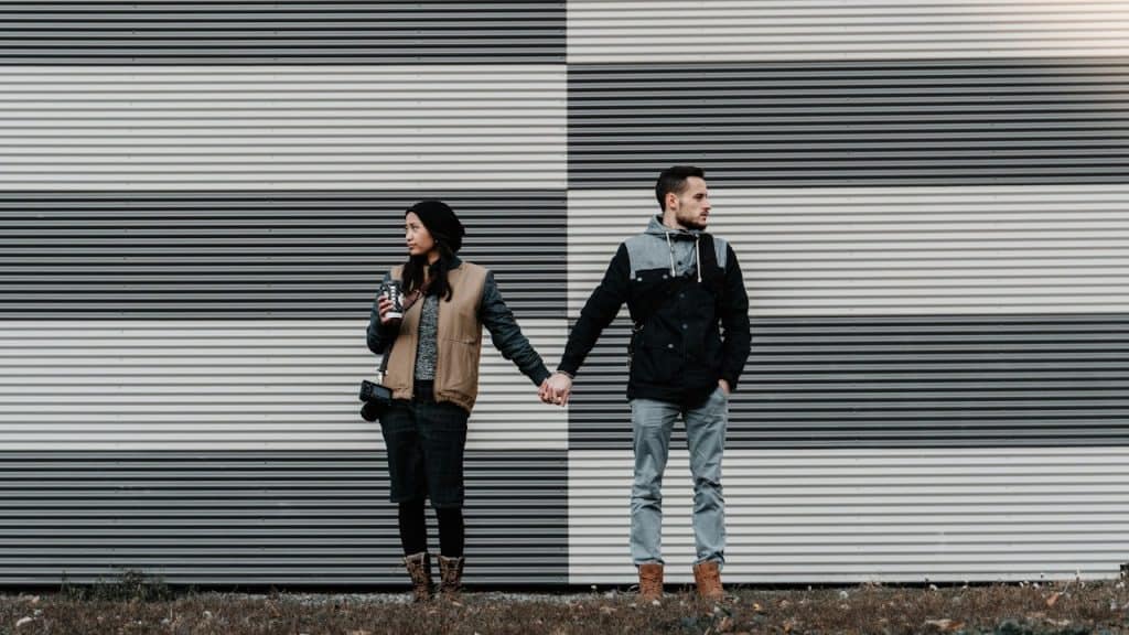 A couple holding hands while standing in front of a corrugated metal wall with alternating horizontal stripes of black and white.