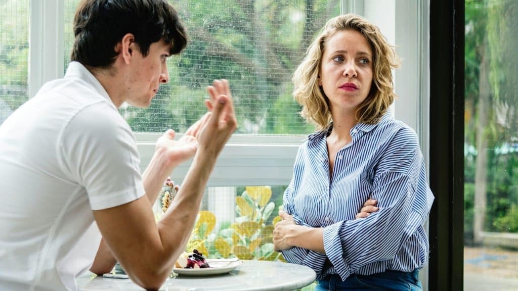 A couple sitting at a table near a window, with the man speaking expressively and the woman looking skeptical with her arms crossed.