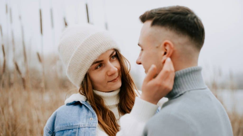 A young woman in a white beanie and a denim jacket affectionately touches the cheek of a man in a gray turtleneck.
