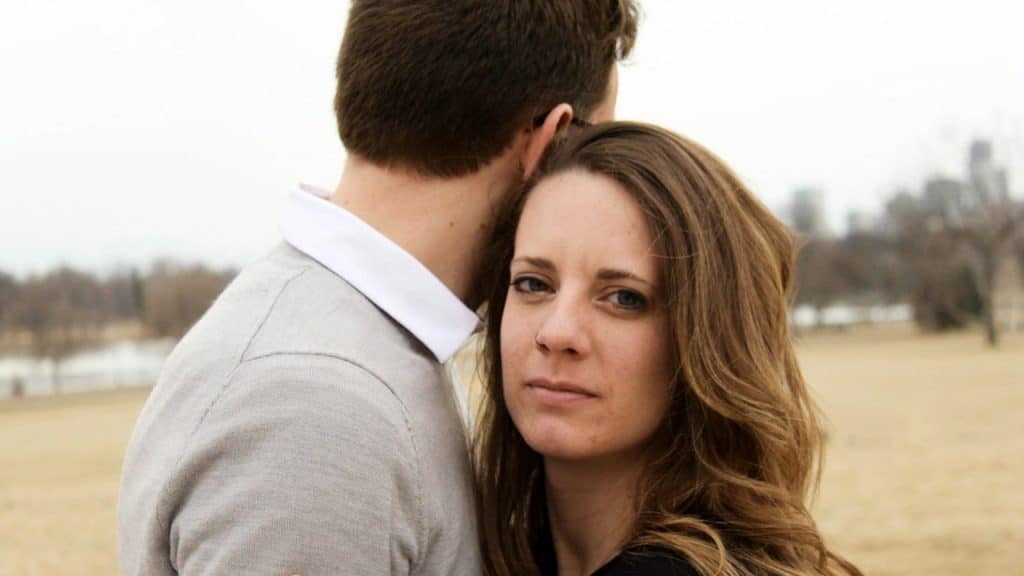 A woman with long wavy hair is looking directly at the camera while embracing a man whose back is turned, standing in a dry, grassy park.