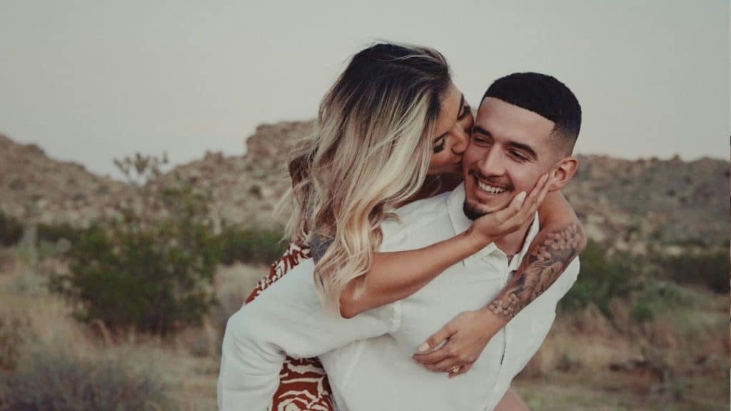 A woman with blonde highlights is giving a man a piggyback ride and kissing his cheek.