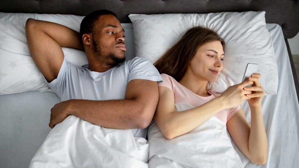 An overhead shot of an interracial couple lying in bed; the woman is smiling while looking at her phone, and the man is looking at her with a jealous expression.