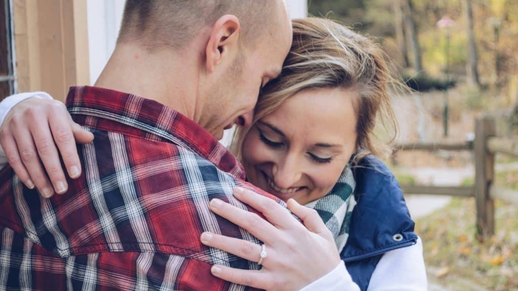 A joyful woman wearing a ring is hugging a man in a red and black plaid shirt, with her eyes closed and a wide smile.
