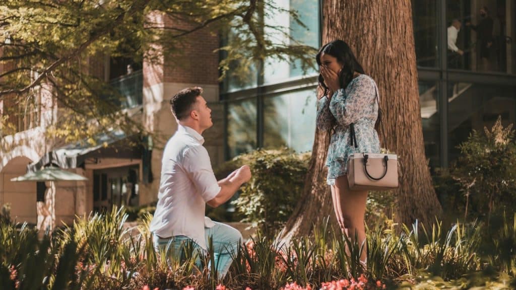 A man in a white shirt is kneeling to propose to a surprised woman in a floral dress.