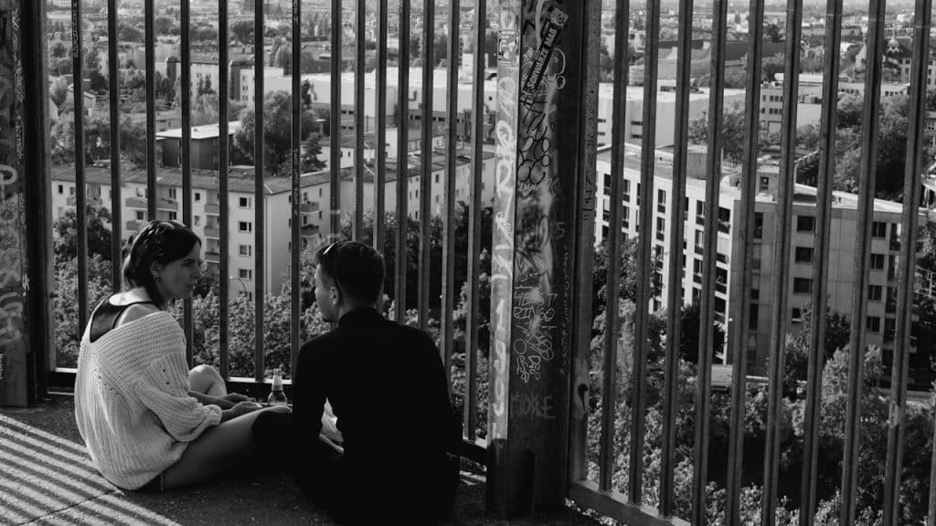 Black and white image of a couple sitting on a high platform, looking out at a cityscape through a metal railing.