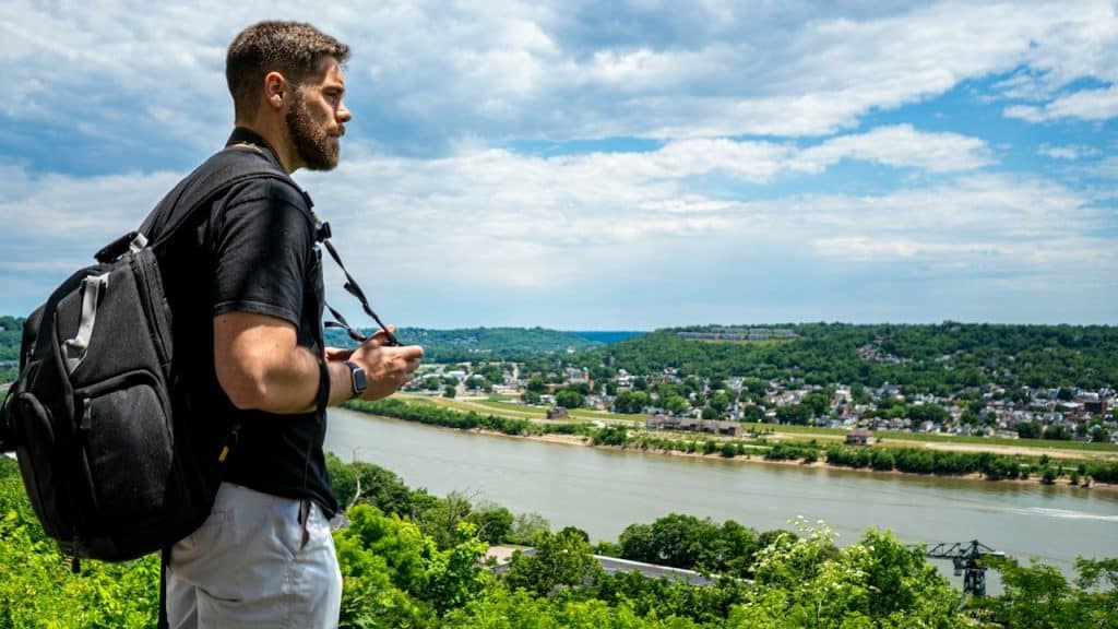 A man in a black t-shirt standing on a rock looking at the city during daytime.