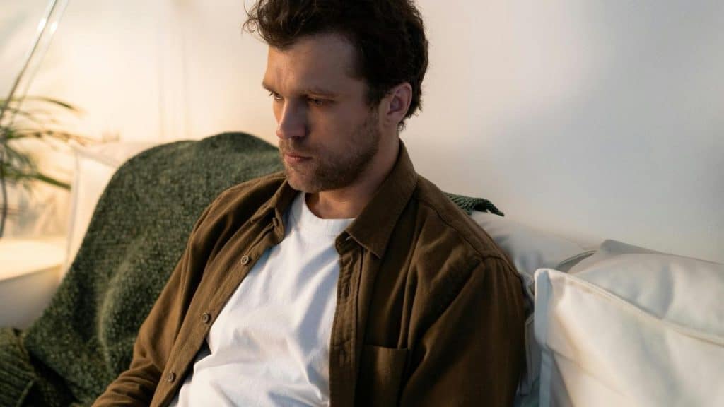 A man sitting on a couch in a softly lit room, appearing deep in thought.