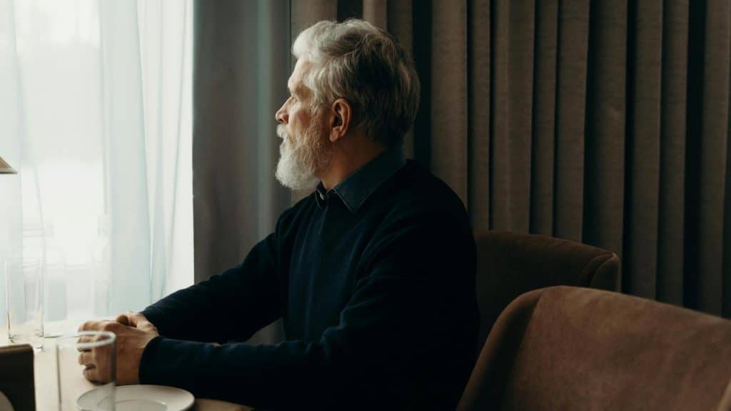 An older man sitting alone at a table, gazing thoughtfully out the window.