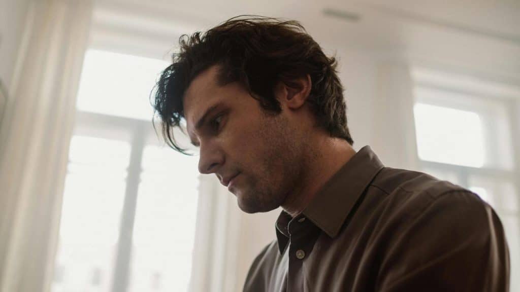 A man in a brown shirt looking down with a thoughtful expression near a bright window.