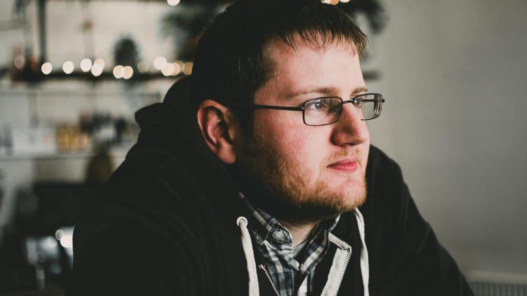 A man wearing glasses and a hoodie sitting indoors, looking off thoughtfully into the distance.