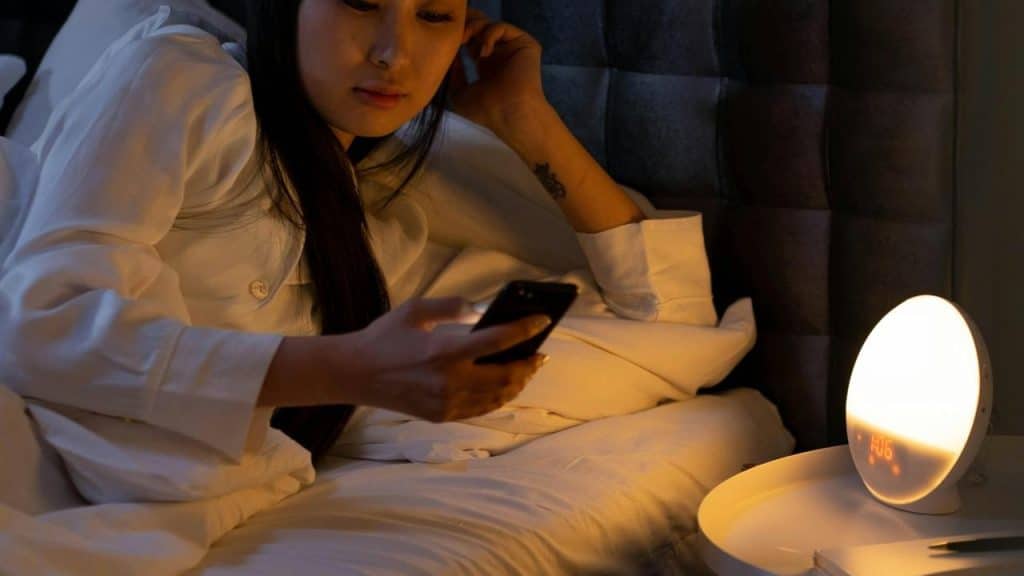A woman in bed wearing white pajamas and looking at her phone beside a lit bedside lamp.