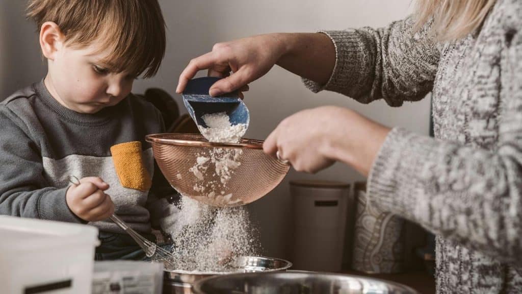 An adult and a child sifting flour together in a kitchen.