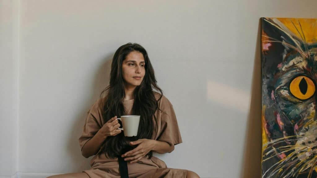 A woman sitting on the floor holding a mug beside a colorful cat painting.