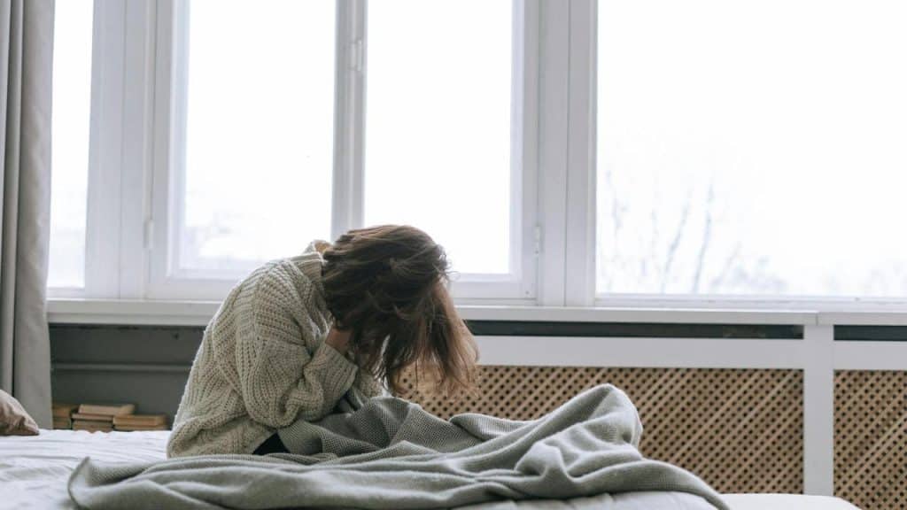 A woman sitting on a bed with her head bowed and hair covering her face.