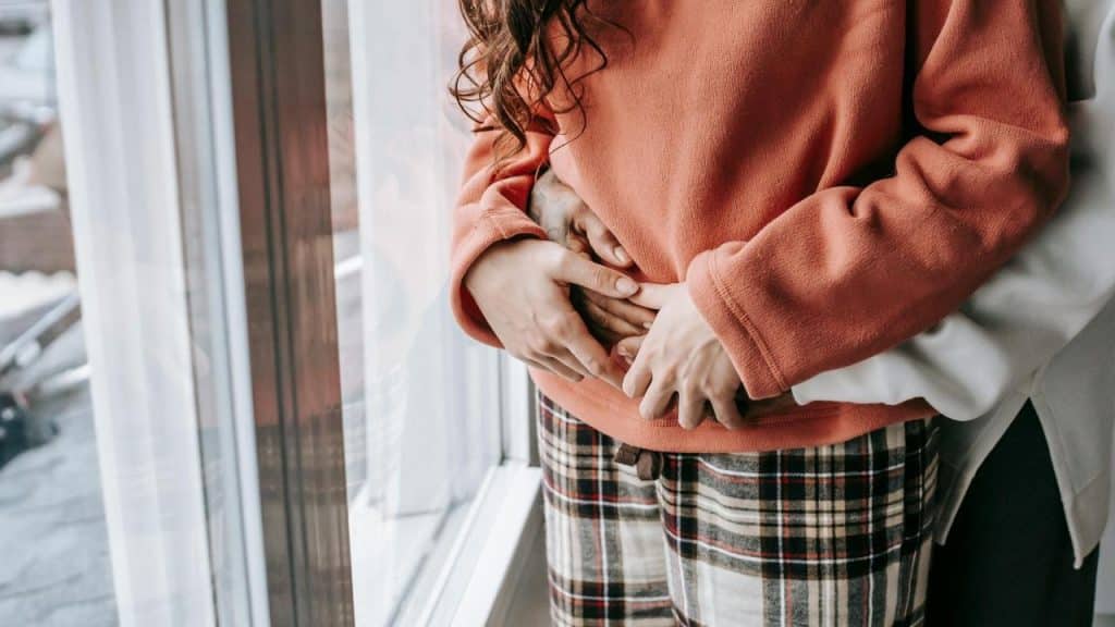 A person in a rust-colored sweater being hugged from behind near a window.