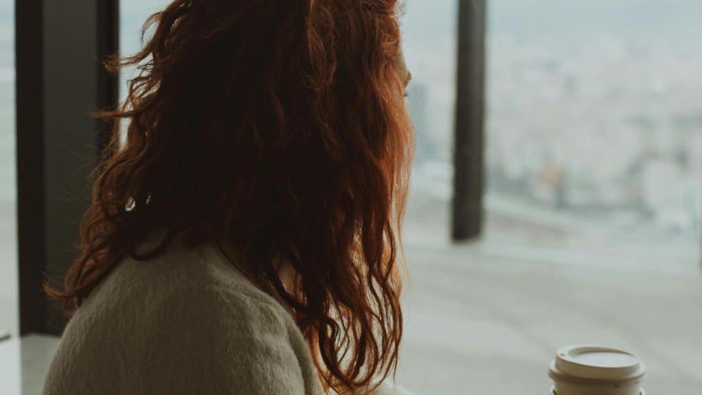 A person with wavy red hair looking out a window while holding a coffee cup.