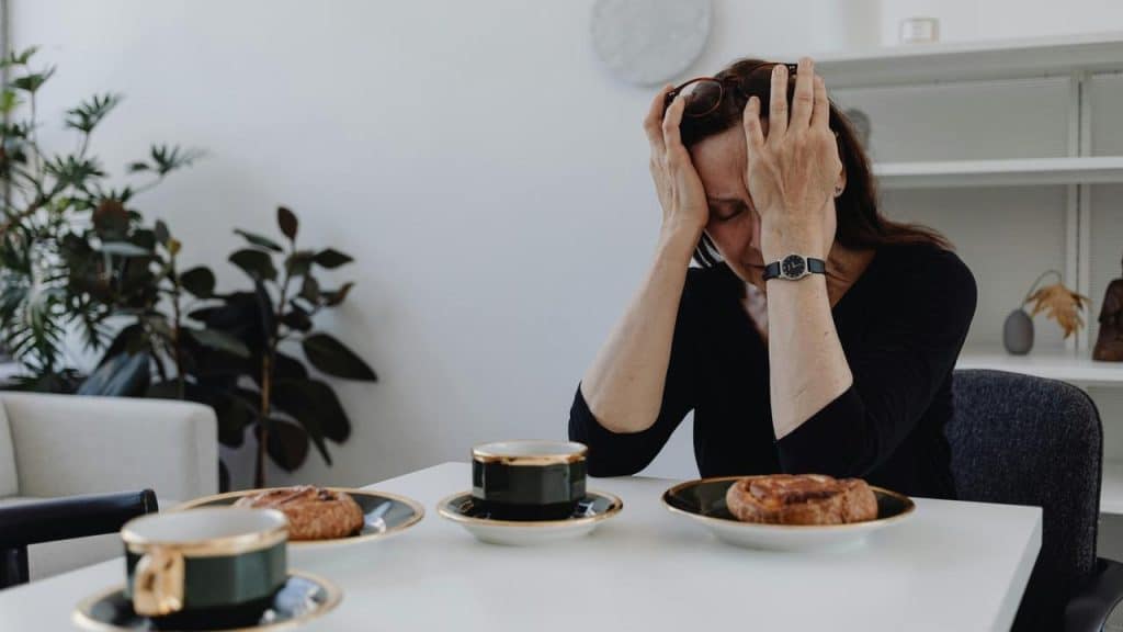 A person sitting at a table with pastries and coffee, holding their head in stress.