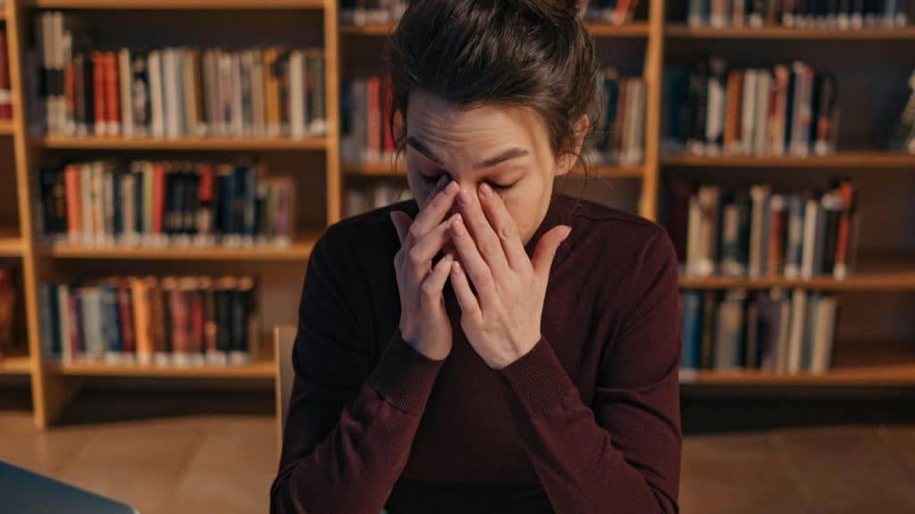 A person sitting in a library rubbing their eyes in frustration or fatigue.