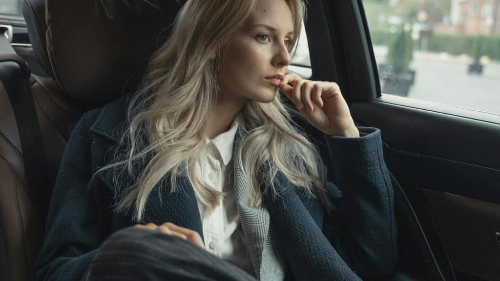 A person with long blonde hair sitting in a car and looking out the window thoughtfully.