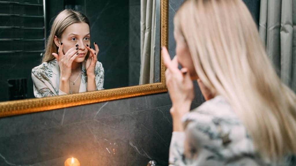 A person standing in front of a mirror, touching their face while looking at their reflection.