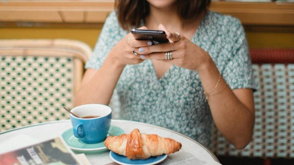 A person sitting at a café table using a phone with coffee and a croissant.
