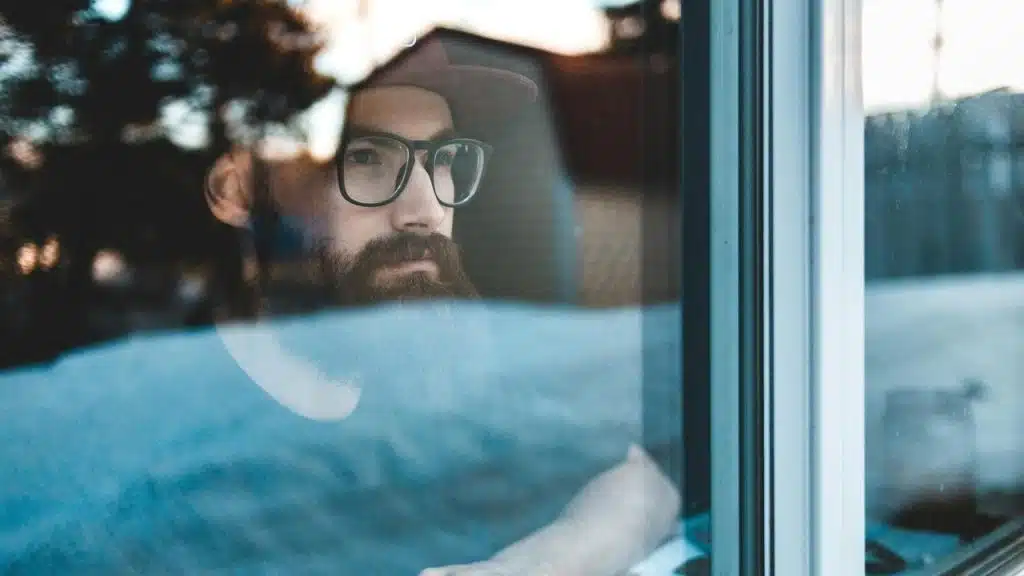 A person wearing glasses and a cap looking thoughtfully out a window.