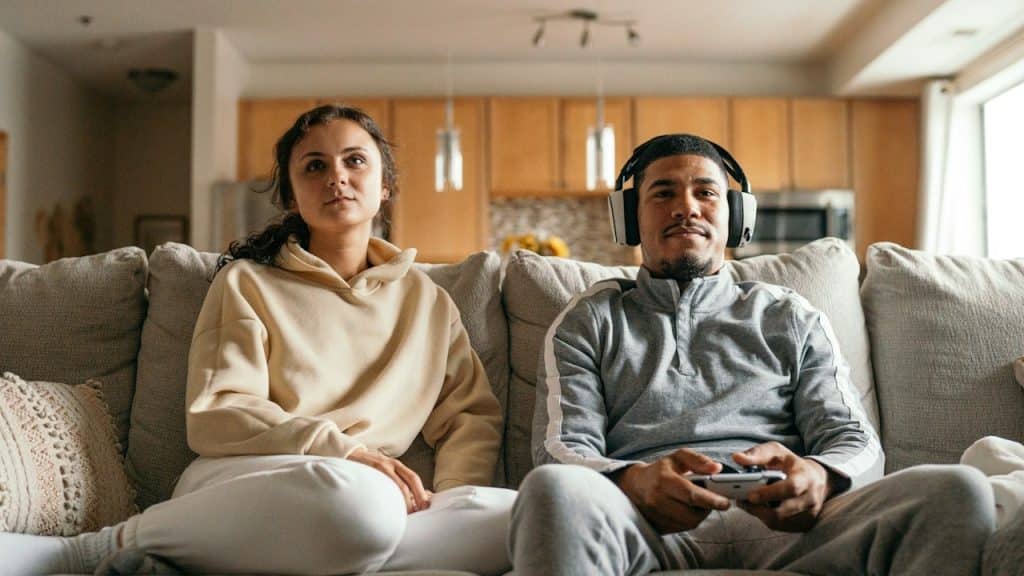 A man is playing a game while holding a controller and wearing a headphone and his wife is sitting with him.