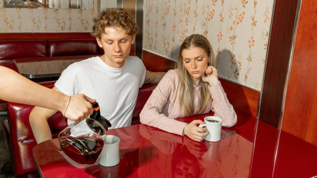 A waiter pours coffee in a mug while a couple is watching silently.