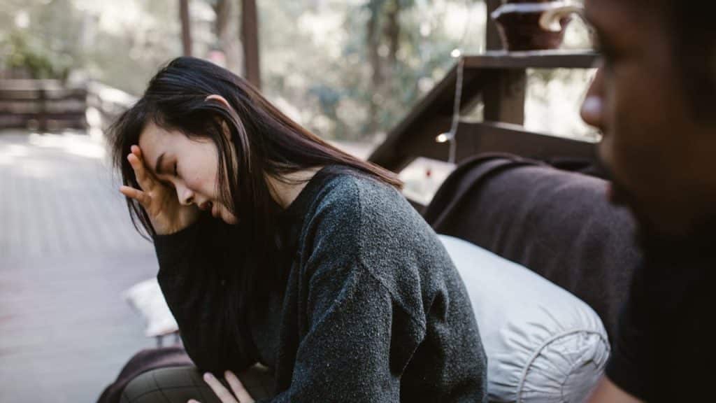 A woman sitting outside with her hand on her forehead, appearing stressed or upset, with a blurred man in the foreground.