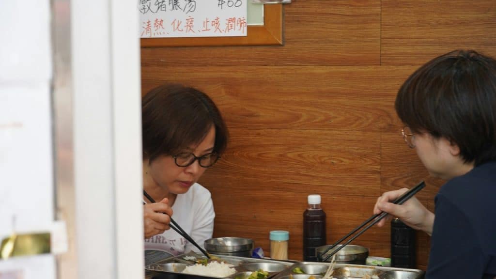 Two people eating with chopsticks out of metal trays at a simple food stall with a wooden wall and a menu sign with Chinese text.