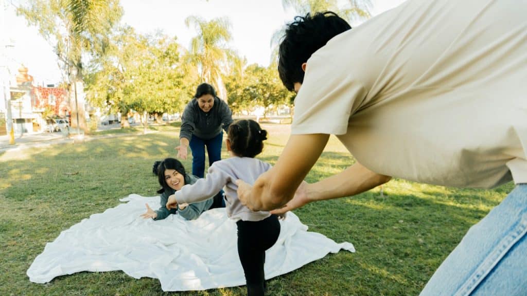 A father holding a toddler's hands as the child takes a step towards two women smiling and lying on a white blanket in a sunny park.