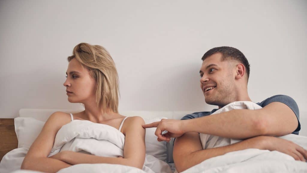 A blonde woman with her arms crossed is looking away from a man who is smiling and trying to get her attention by poking her back in bed.