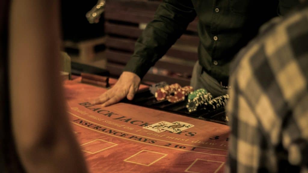 A close-up of a casino table with a dealer's hand near the Blackjack felt, showing a few cards and stacks of poker chips.