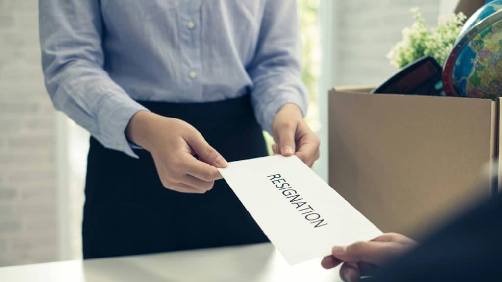 A person in a blue shirt is handing a resignation letter to another person across a white desk, with a cardboard box full of personal items in the background.