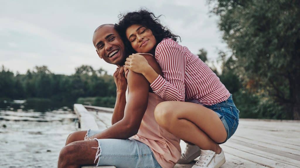 A happy young couple embracing and smiling while sitting on the pier near the lake.