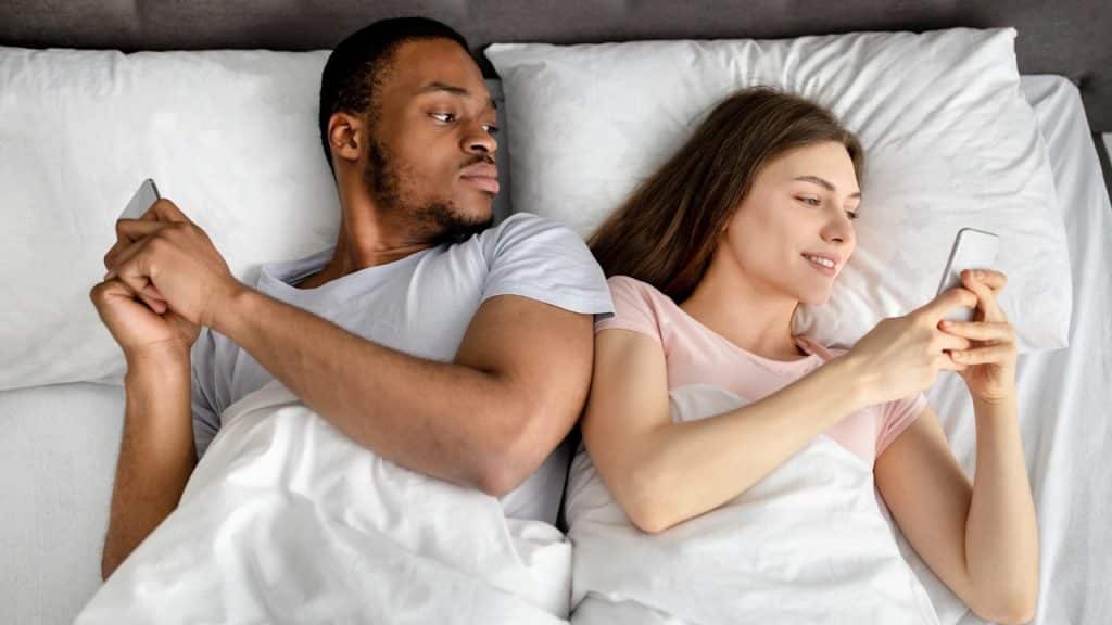 An overhead shot of a couple in bed, both looking at their smartphones, with the man looking suspiciously at the woman's phone.