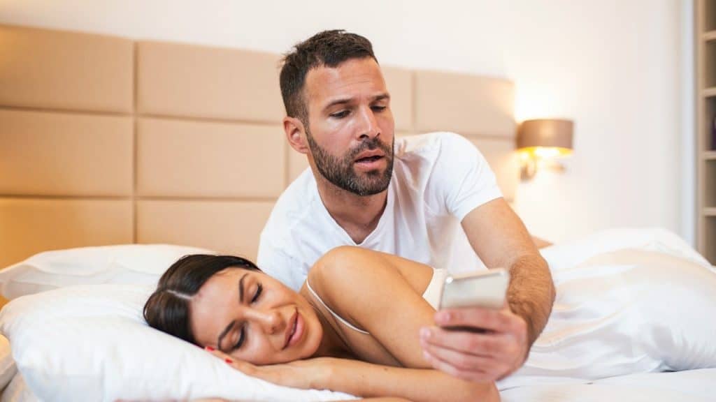 A surprised-looking man leans over a woman, who is lying on her side, to look at her smartphone while they are in bed.