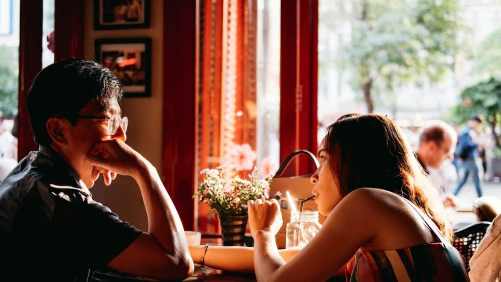 A woman sipping on a straw is watching a man wearing a glasses who is looking towards her.
