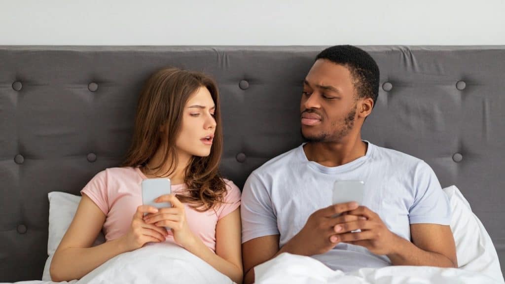 A woman and a man are sitting in bed, both looking at their smartphones and glancing suspiciously at each other.