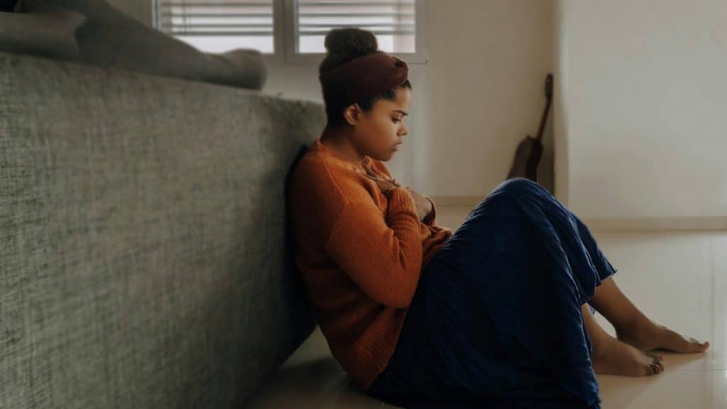 A barefoot woman sitting on a tiled floor with her back against a sofa, holding her chest