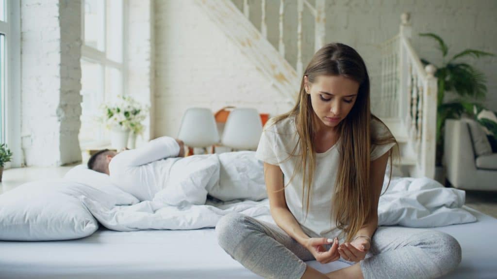 A sad woman is sitting cross-legged on a bright white bed, while a man is sleeping under the covers behind her